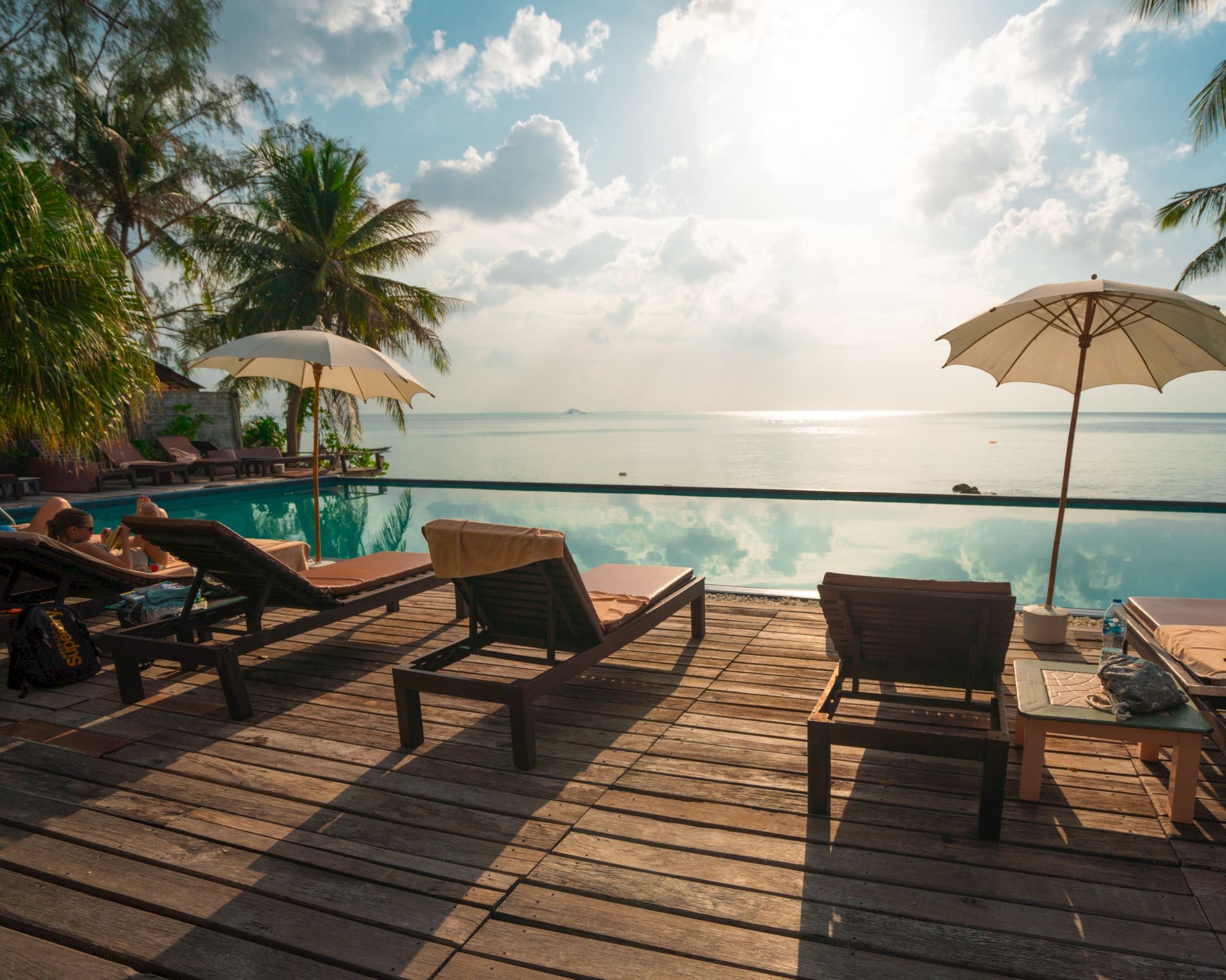 A tropical beachfront resort scene with loungers and umbrellas beside a pool, overlooking the ocean under sunny clouds.
