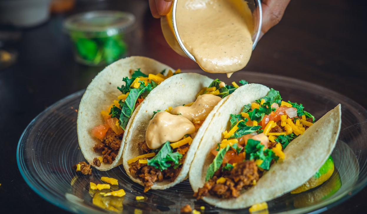 Three tacos with shredded meat and colorful toppings as creamy sauce is drizzled on top, ready to eat.
