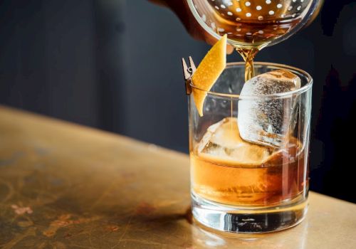 A whiskey glass with ice and an orange twist, being poured from a bottle, on a wooden bar top. End.