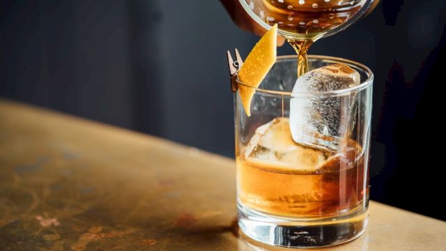 A whiskey glass with ice and an orange twist, being poured from a bottle, on a wooden bar top. End.