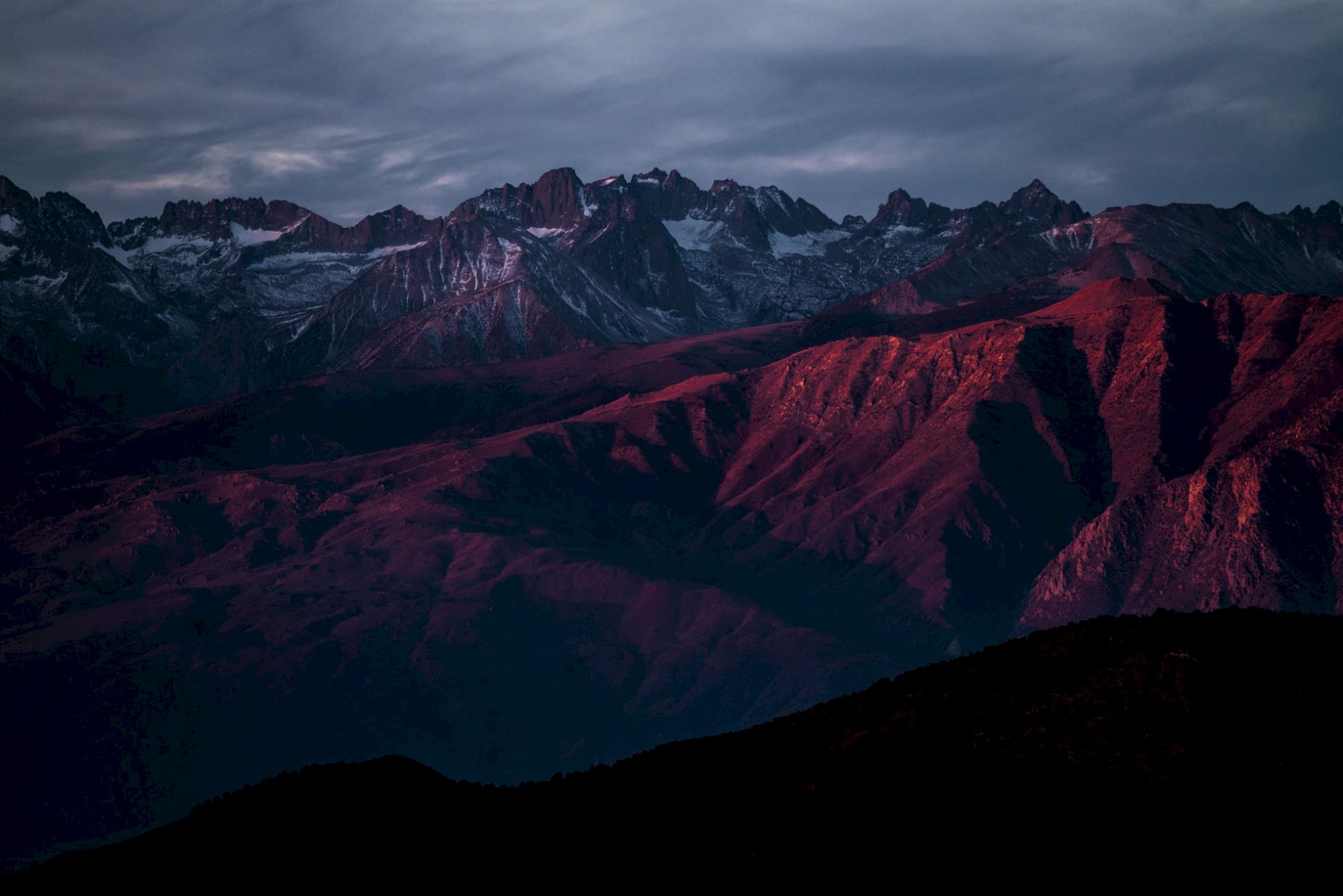 A dramatic mountain range at dusk, with snow patches and reddish light glowing across rocky peaks and valleys under dark clouds.