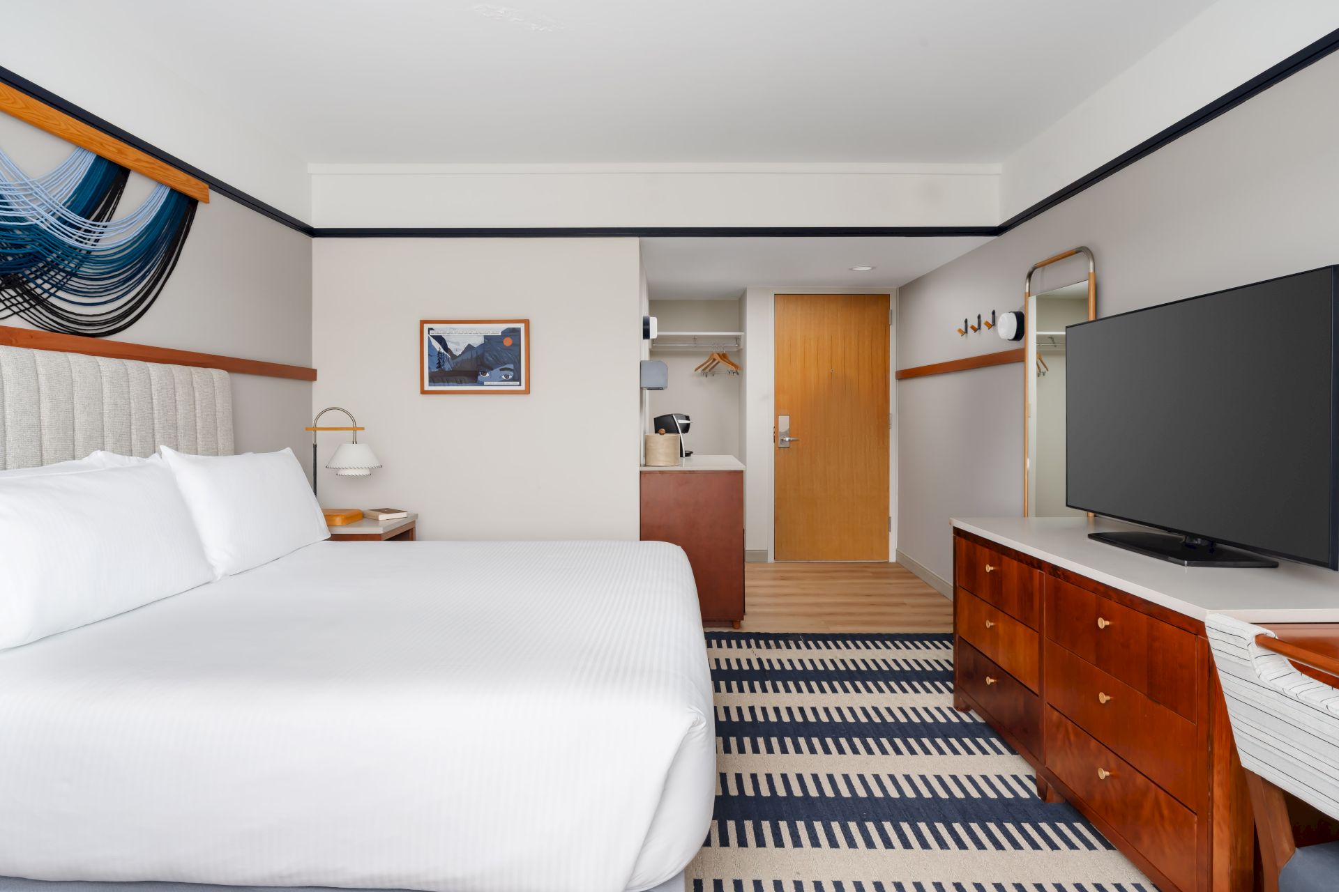 A modern hotel room with a large white bed, wooden dresser, flat-screen TV, striped rug, and a small desk area near an open wardrobe.