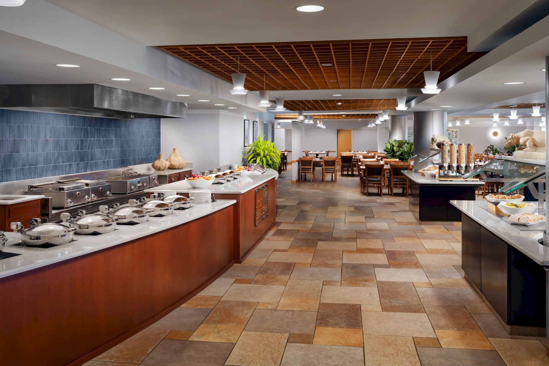 A bright hotel buffet kitchen with stainless cooking stations, serving counters, plants, and warm overhead lights.