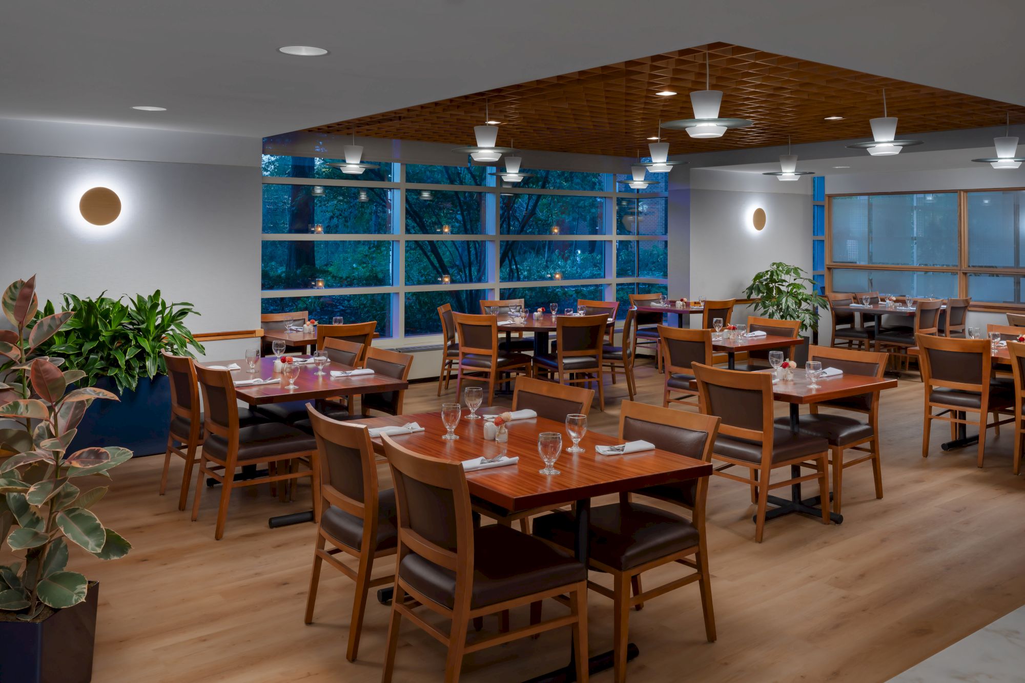 A bright restaurant dining room with wooden tables and chairs, place settings, plants, and large windows, creating a warm, modern atmosphere.