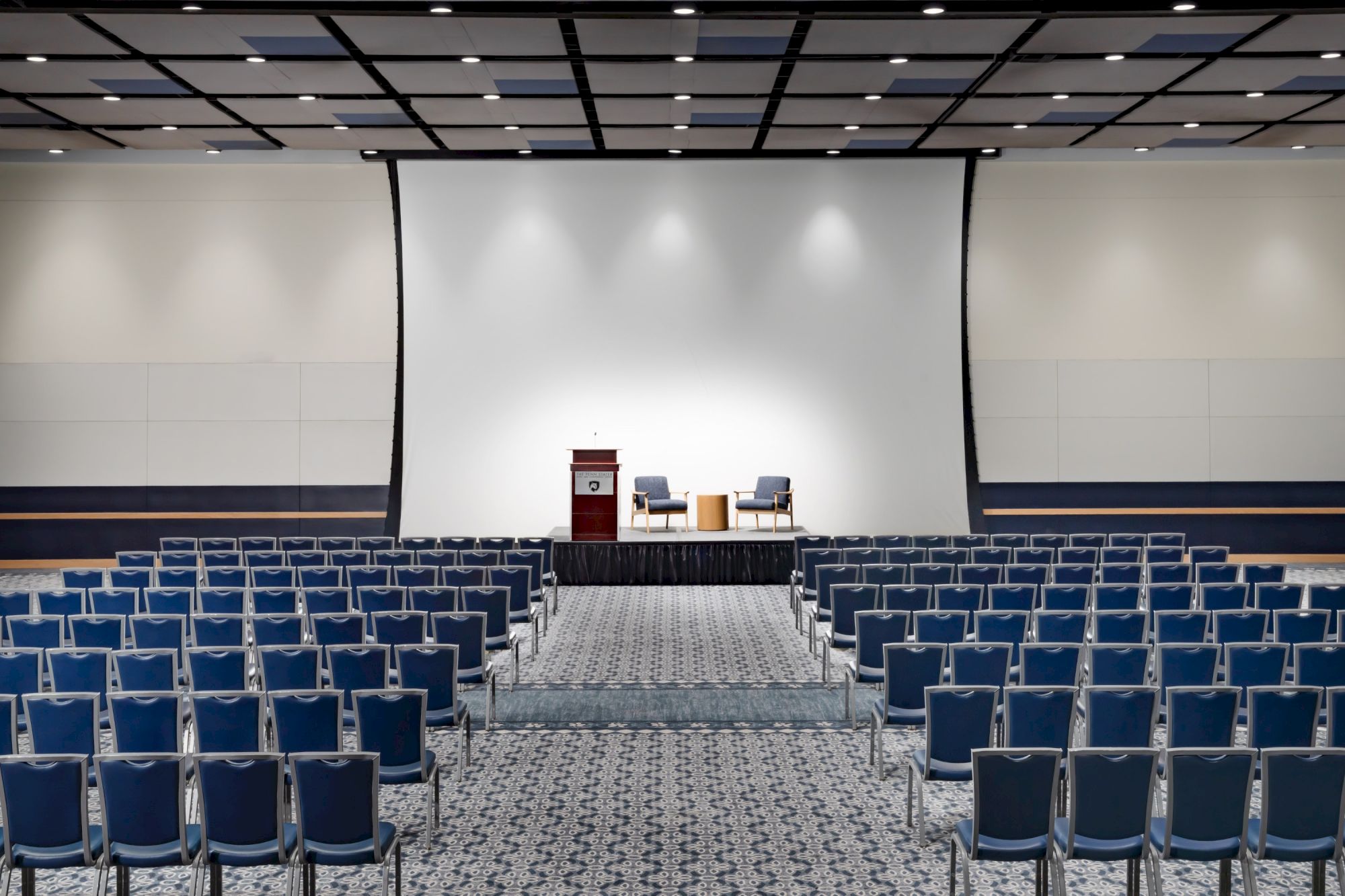A conference or seminar setup with a large stage, podium, and two chairs, surrounded by hundreds of blue-cushioned chairs facing the stage.