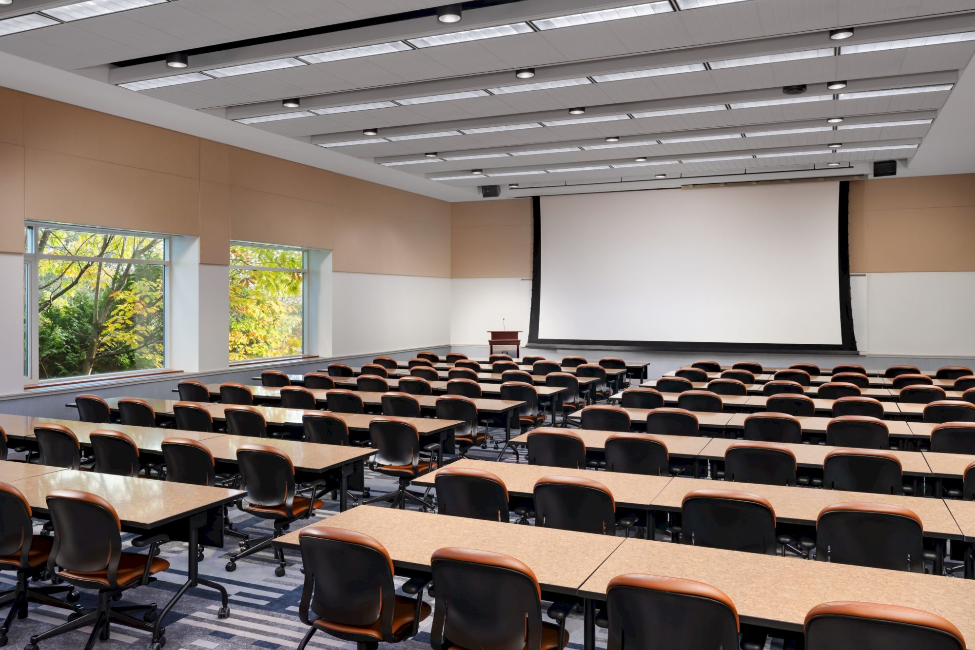 A large classroom or lecture hall with long rows of desks and chairs facing a big projector screen at the front, windows on the side, and a modern ceiling.