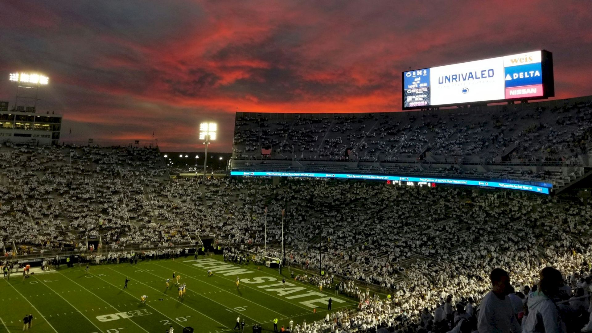 A crowded football stadium at dusk with a glowing sunset, players on the field, and a large scoreboard in the distance.
