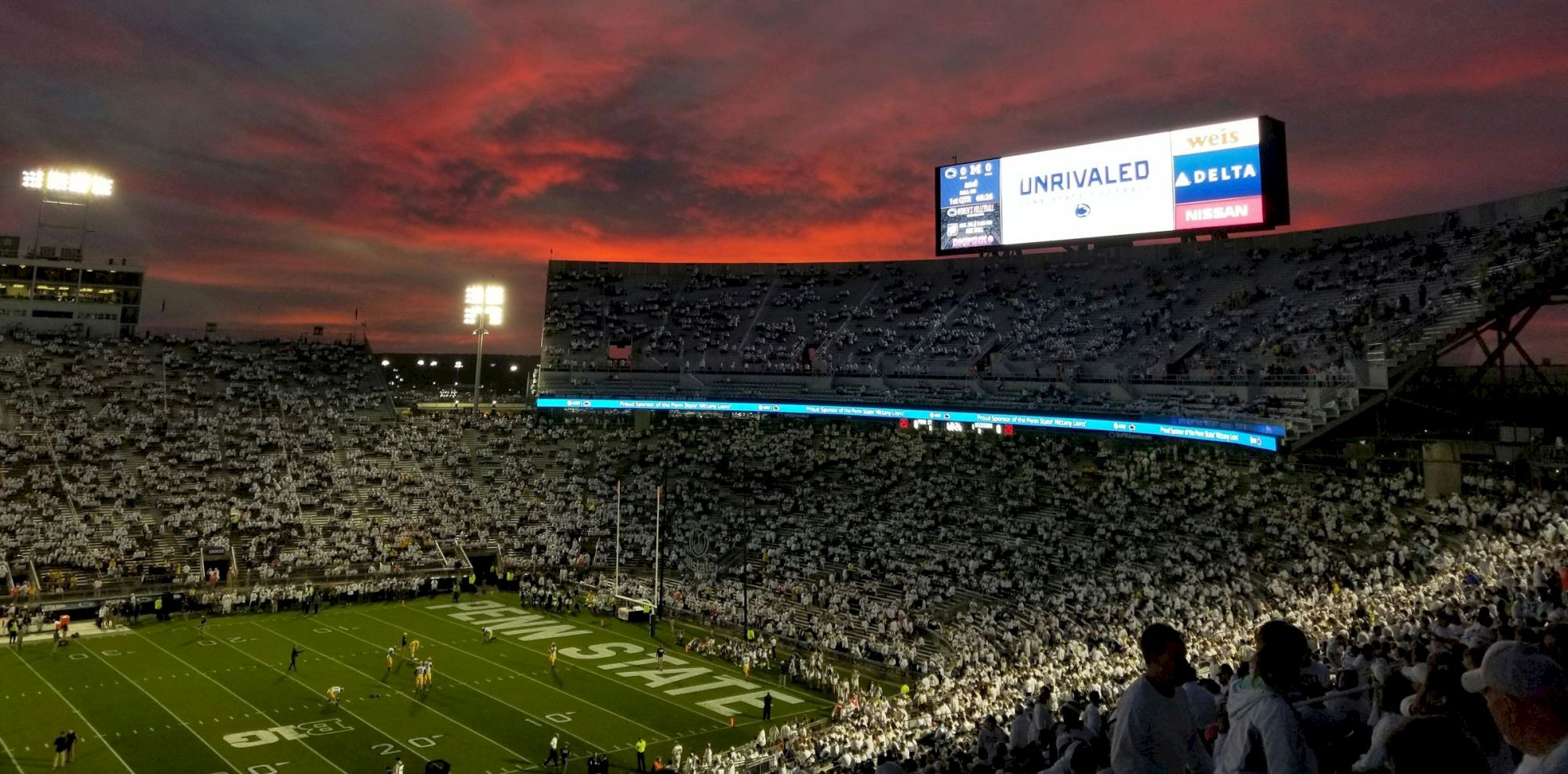A crowded football stadium at dusk with a glowing sunset, players on the field, and a large scoreboard in the distance.