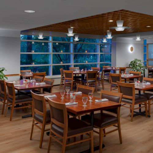 A modern dining area with wooden tables and chairs, wine glasses, pendant lighting, potted plants, and large windows, tidy and inviting.