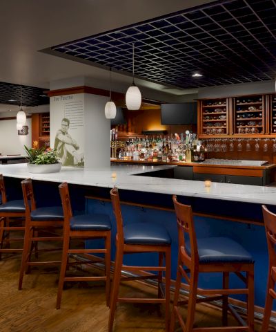 A sleek, empty bar with a long marble counter, blue stools, warm lighting, shelves of bottles, and a modern lounge vibe, ready for guests.