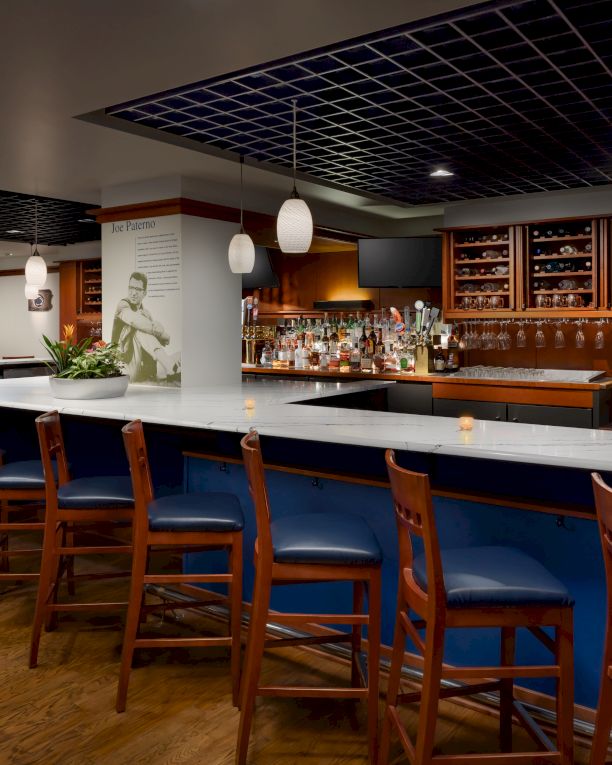 A sleek, empty bar with a long marble counter, blue stools, warm lighting, shelves of bottles, and a modern lounge vibe, ready for guests.