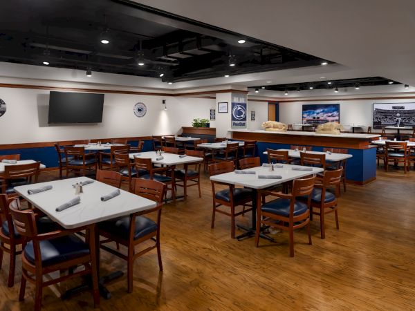 A large, empty restaurant or dining hall with neatly set tables and chairs, wood floors, and flat-screen TVs on the walls, ready for guests.
