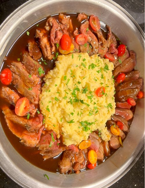 Succulent roast beef with a rich brown sauce, surrounding a fluffy mound of mashed potatoes garnished with chopped herbs and colorful vegetables, all plated neatly.
