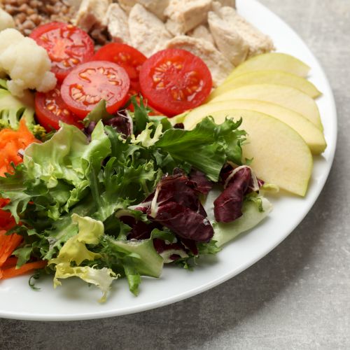 Plate with a mixed salad: cherry tomatoes, sliced apples, shredded carrots, leafy greens, cucumber/cauliflower florets, and diced chicken on a white plate.