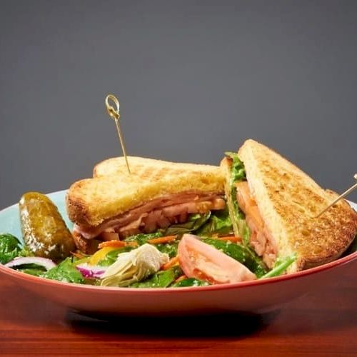 A plated sandwich with toasted bread, greens, and pink fillings, skewered tips and a side salad on a shallow bowl.