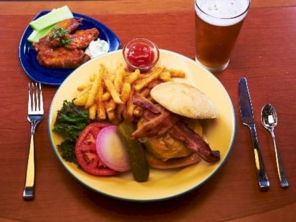 A hearty plate with a burger, bacon, fries, greens, tomato, onion, pickle, and a side of dipping sauce, plus a beer on the side.