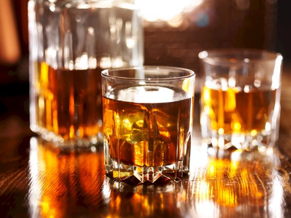 A clear rock-glass filled with amber whiskey sits on a wooden bar, with warm bottles blurred in the background.