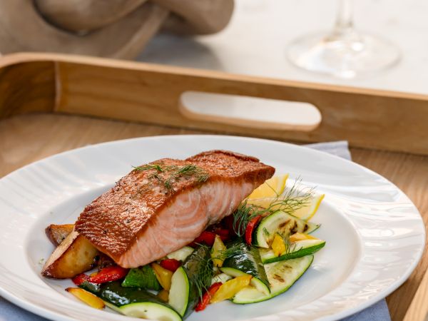 A plated salmon fillet on a vegetable medley, served on a white dish with a wooden tray, wine glass in the background, and a knotted rope in view.