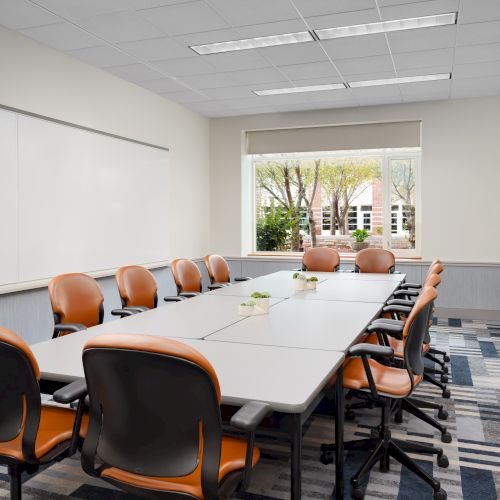 A modern conference room with a long table, orange-and-black chairs, whiteboards, and a window view of trees outside, ready for a meeting.