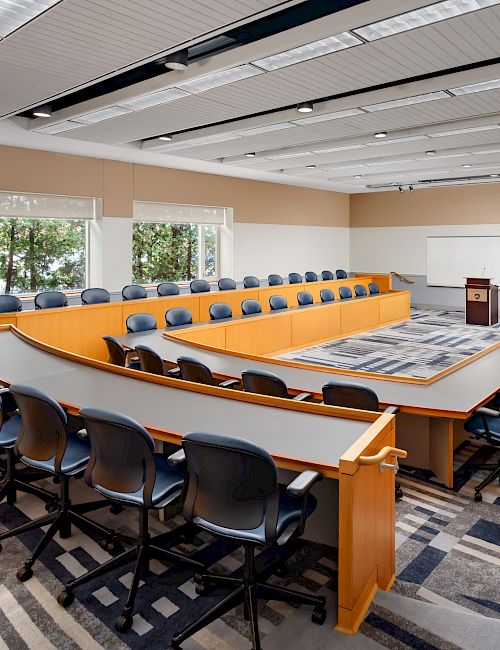 A large U-shaped conference room with brown desks, blue chairs, a podium, and a whiteboard; windows on one side provide natural light.
