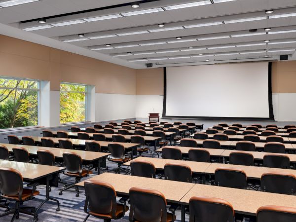 A large empty lecture hall with rows of desks and chairs facing a projector screen, windows on the left letting in light, and a podium at the front.