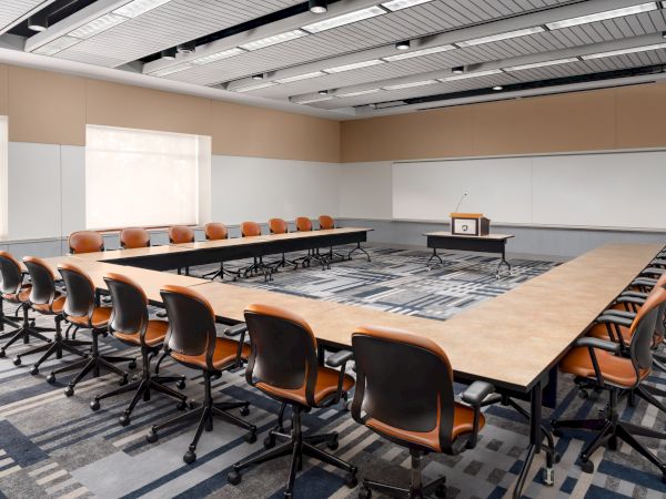 A large conference room with a U-shaped table setup, orange cushioned chairs, whiteboard walls, and a projector at the front.
