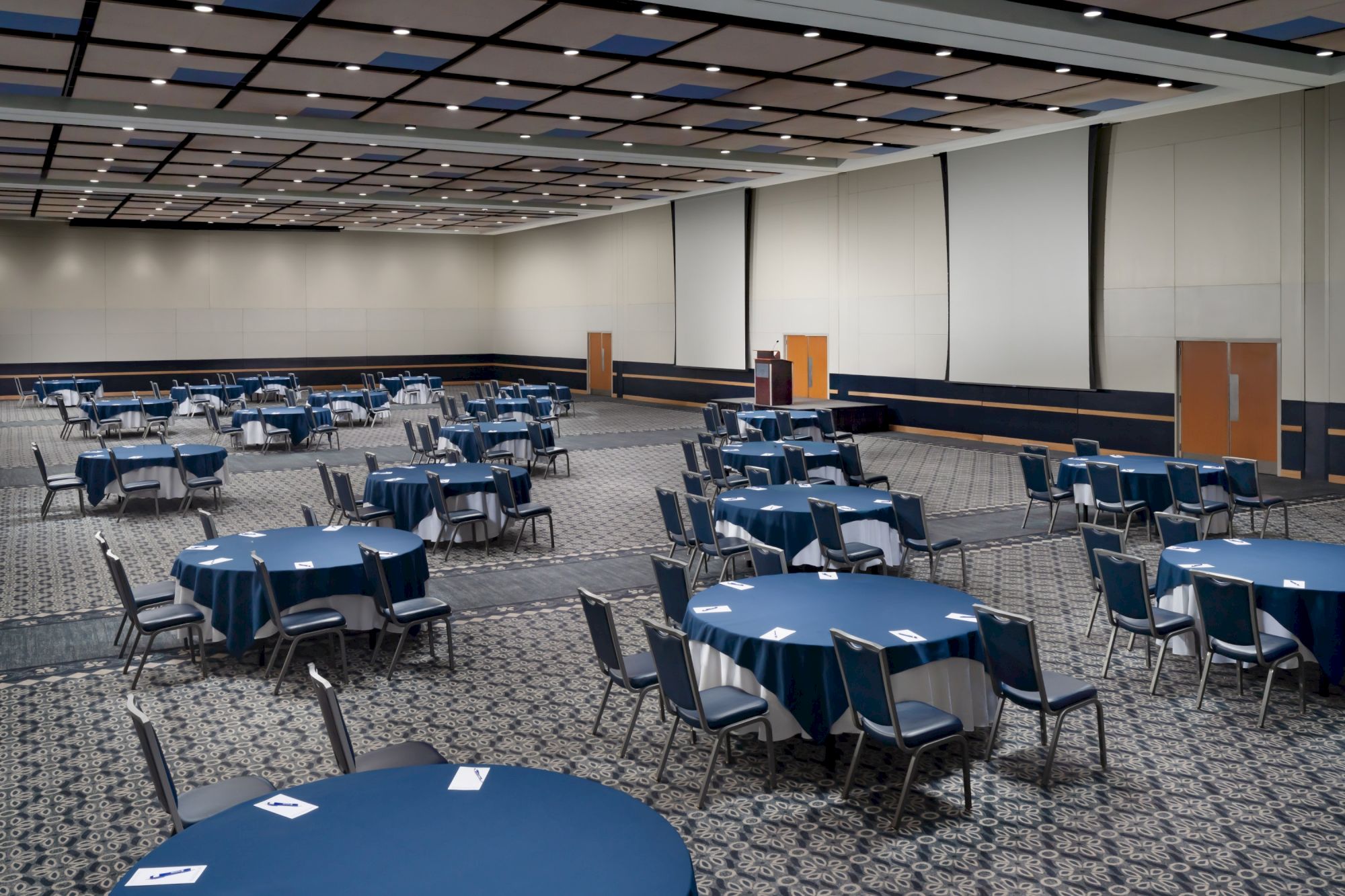A large, empty banquet hall with round blue tables and blue chairs arranged neatly, ready for an event, spacious with ceiling lights.