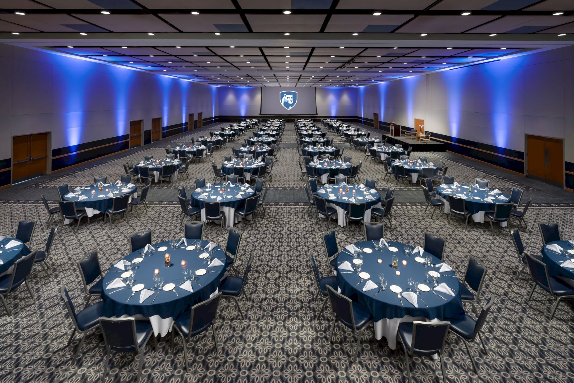 A large banquet hall set for a formal event, with round tables, navy linens, folded napkins, and soft blue uplighting along the walls.
