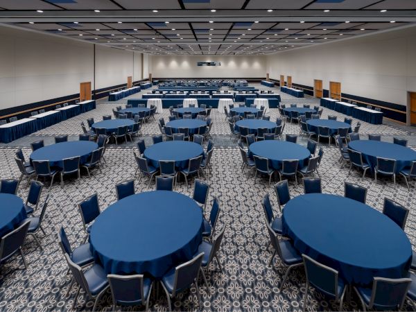 A large banquet-style conference hall with many round tables covered in blue tablecloths and matching chairs, arranged in rows for an event.