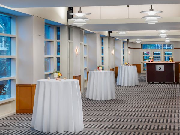 A bright event space with tall round cocktail tables draped in white linens, soft lighting, and a long hallway setup near windows.