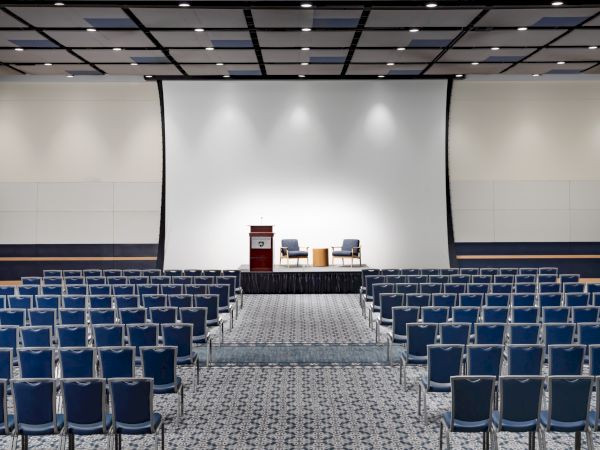 A large conference hall with a stage, podium, and two chairs facing rows of blue seats under bright ceiling lights.