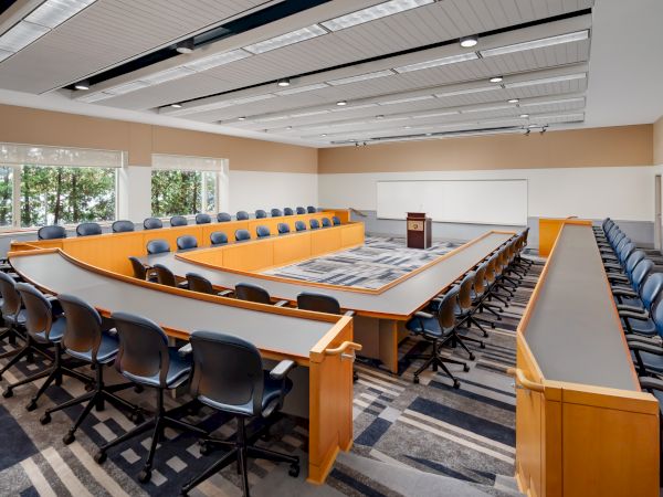 A large, modern conference room with a U-shaped table, orange accents, and many blue chairs; whiteboards, podium, and bright windows showing greenery.