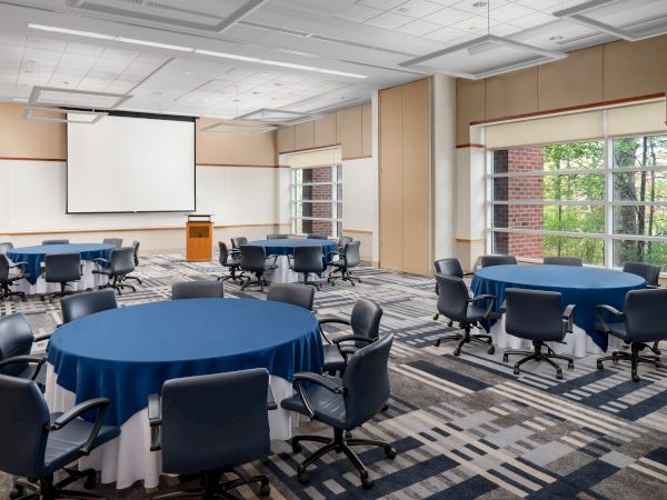 A conference room set for a meeting with round tables draped in blue cloth, black chairs, a projector screen, and large windows.