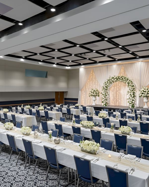A spacious event hall set for a wedding or banquet, with long tables, white linens, blue chairs, floral centerpieces, and a decorated altar backdrop.