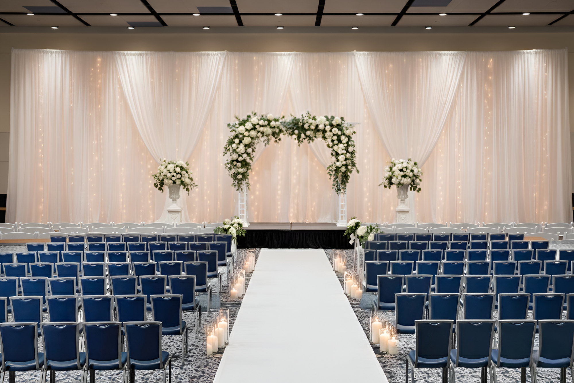 A wedding ceremony setup with a white aisle, floral arch, and draped backdrop, flanked by blue chairs and lanterns along the aisle end. End.