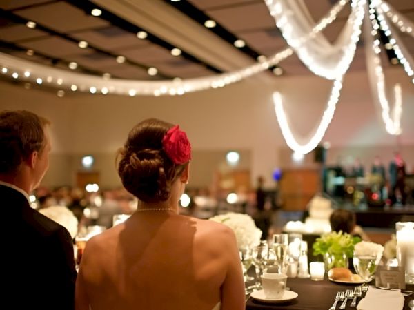 A couple dressed up at a formal reception, seated at a candlelit banquet with string lights overhead, watching a stage or ceremony begin.