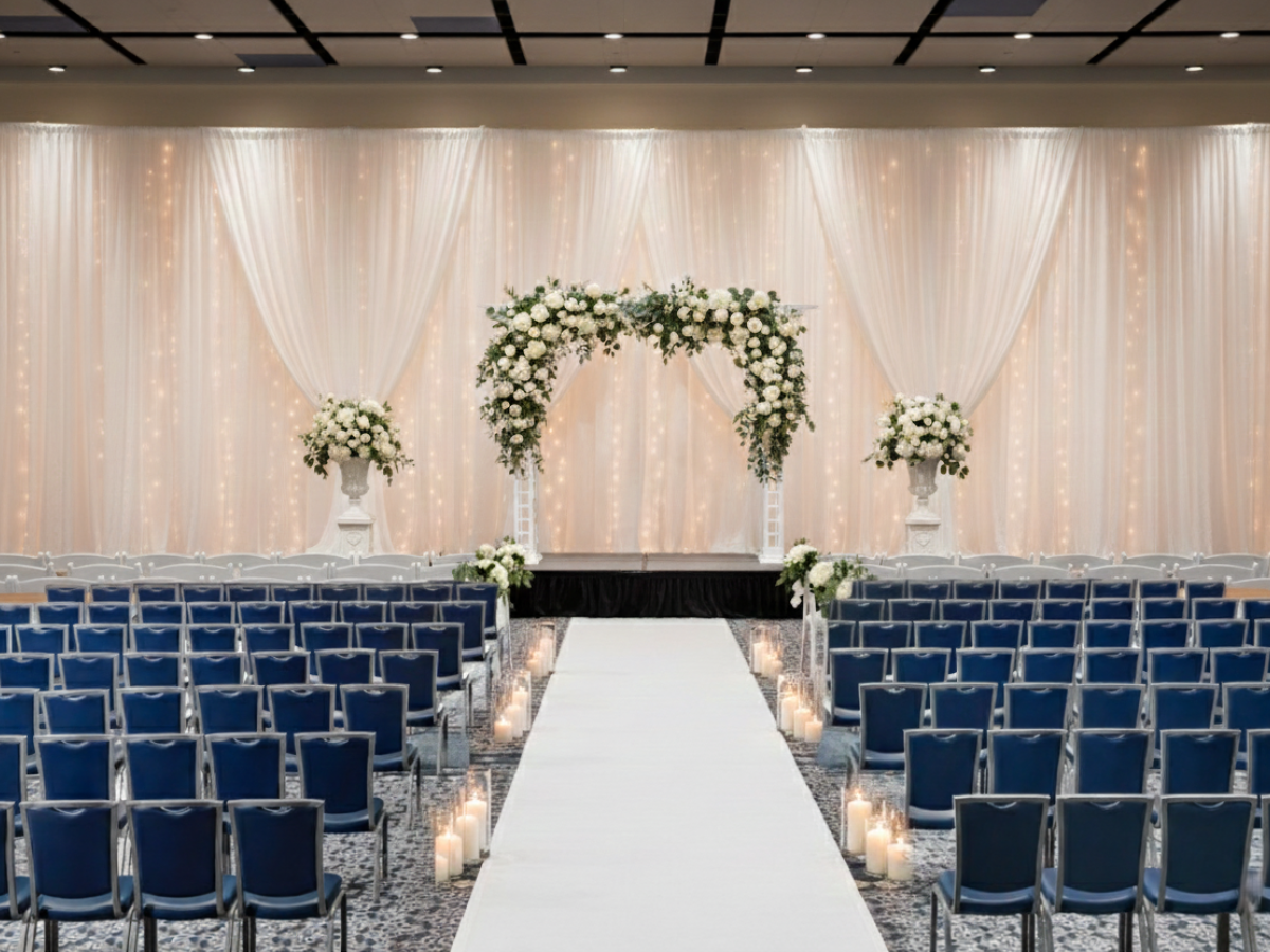 A wedding ceremony setup with a white aisle, blue chairs, and floral archways at the front, softly glowing lights, and draped backdrop.