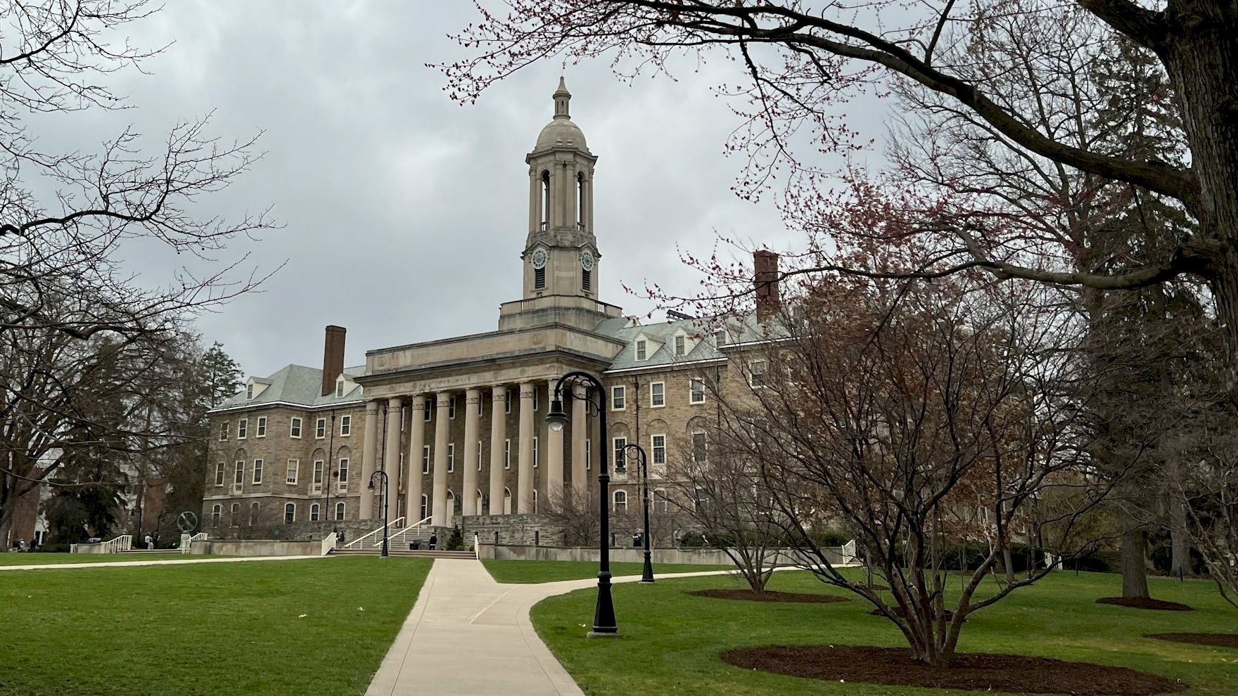 A grand neoclassical building with a central clock tower, surrounded by a manicured lawn, leafless trees, and a paved walkway ahead.