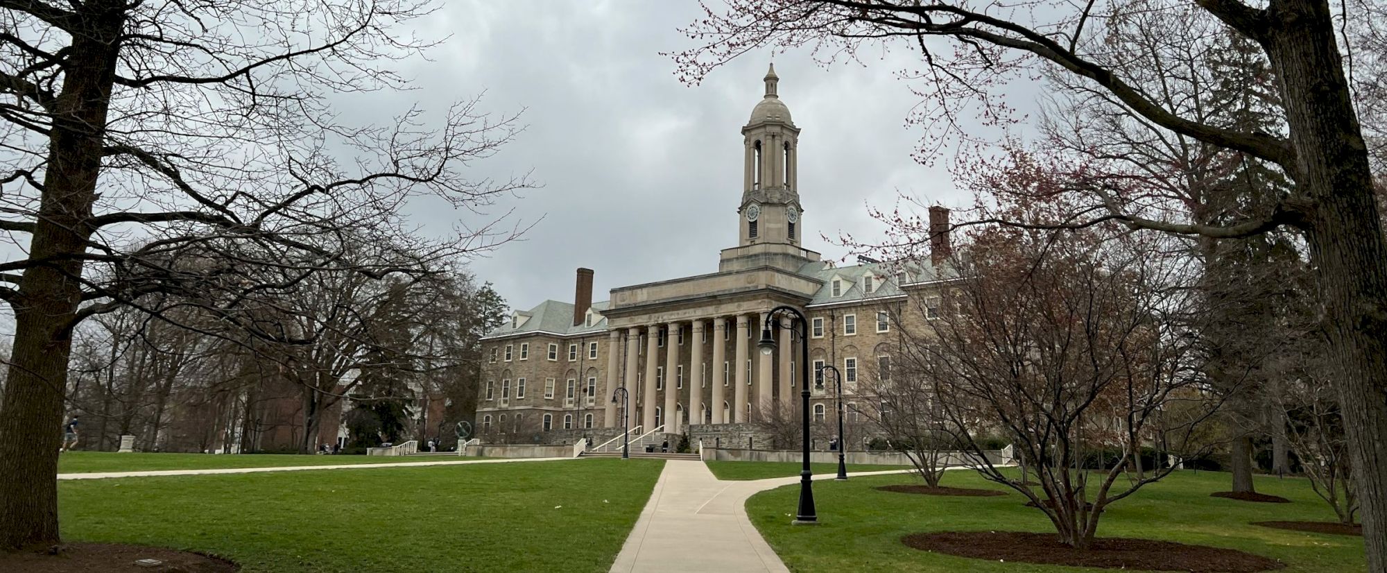 A grand neoclassical building with a central clock tower, surrounded by a manicured lawn, leafless trees, and a paved walkway ahead.