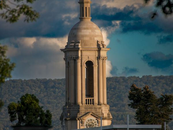 A tall, ornate clock tower or cupola rises above a historic building, framed by trees, with a dramatic blue sky and distant hills in the background.