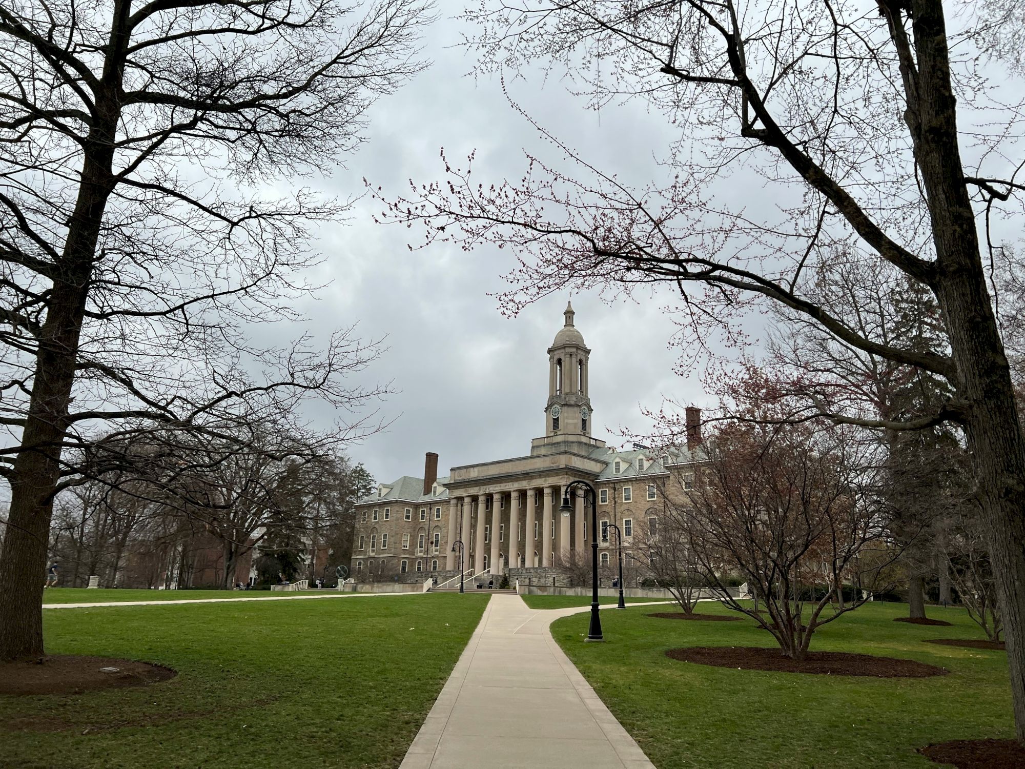 A large neoclassical building with a central tower sits beyond a paved walkway, framed by leafless trees on a grassy campus.