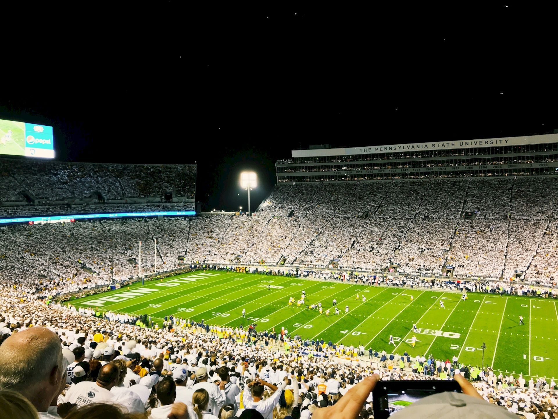 A packed football stadium at night, white-clad fans fill the stands while the illuminated field hosts a game under bright lights.