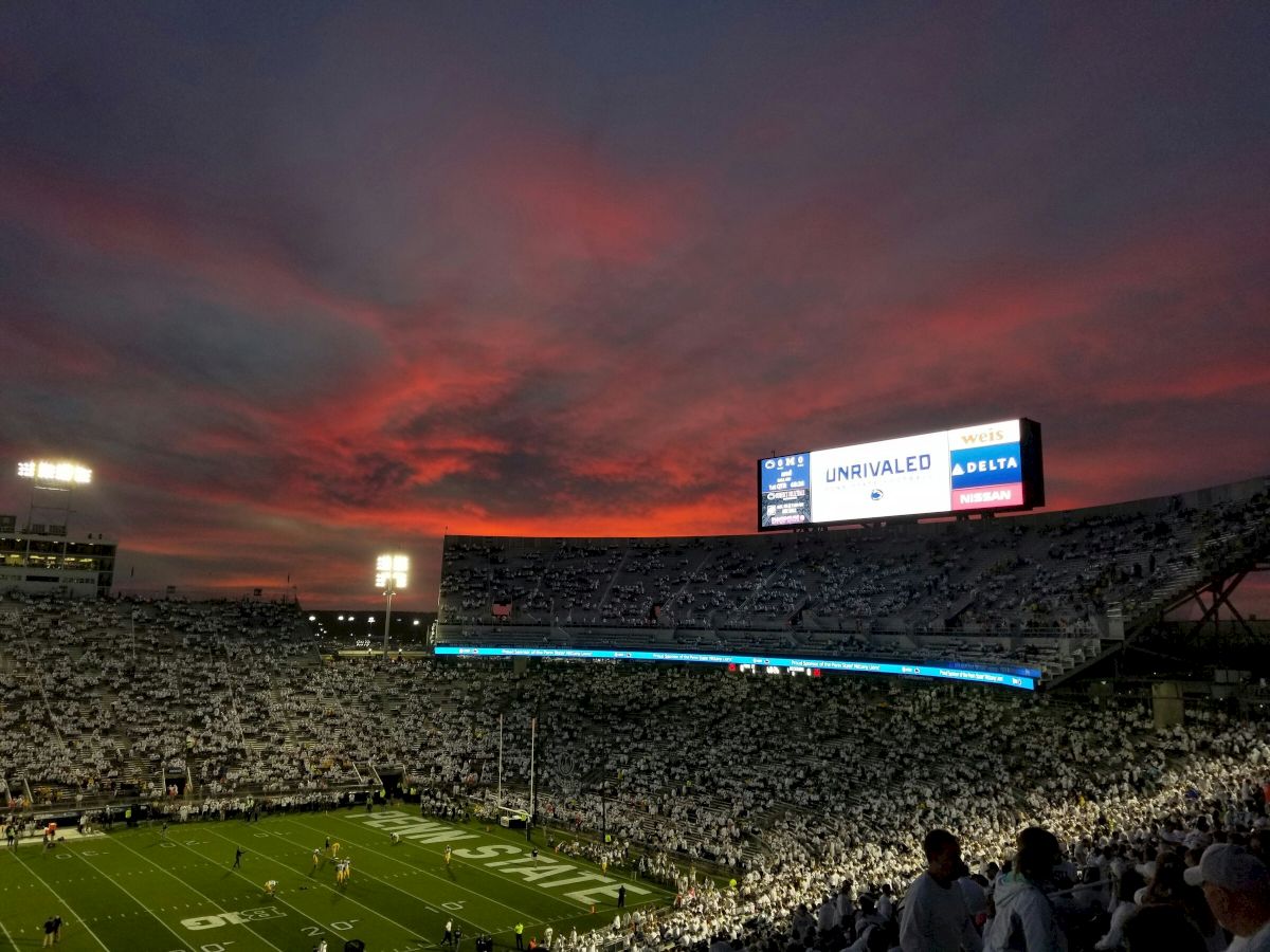 A packed football stadium at dusk, red-orange sunset sky, players on the field, and bright stadium lights illuminating the stands.