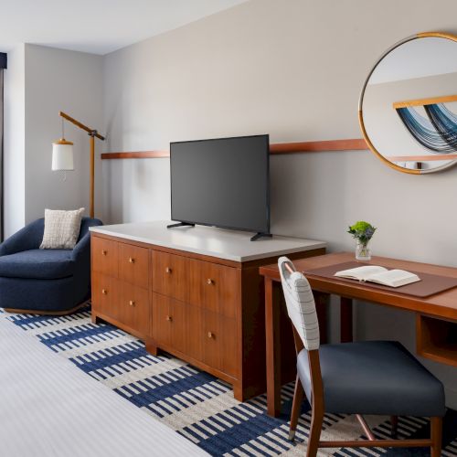 A hotel room with a TV on a wooden cabinet, a desk and chair, a round mirror, a blue chair, rug, and a window with curtains. Top it at 140 characters, always ending the sentence.