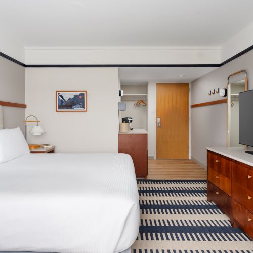 A bright hotel room with two large white beds, a striped rug, flat-screen TV on a wooden dresser, and a doorway to a small kitchenette area.