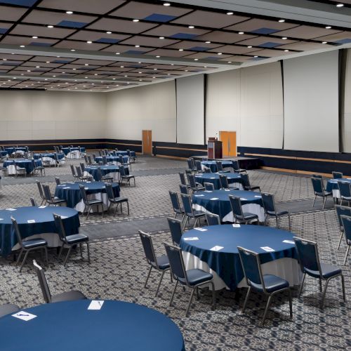 A large conference room set up with multiple round blue tables and chairs, spaced out across a long hall with a patterned carpet and white walls.