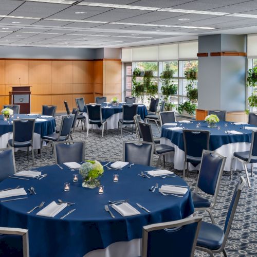 A spacious conference room set for a meal, with round tables covered in blue tablecloths, neat place settings, and plants by the windows.