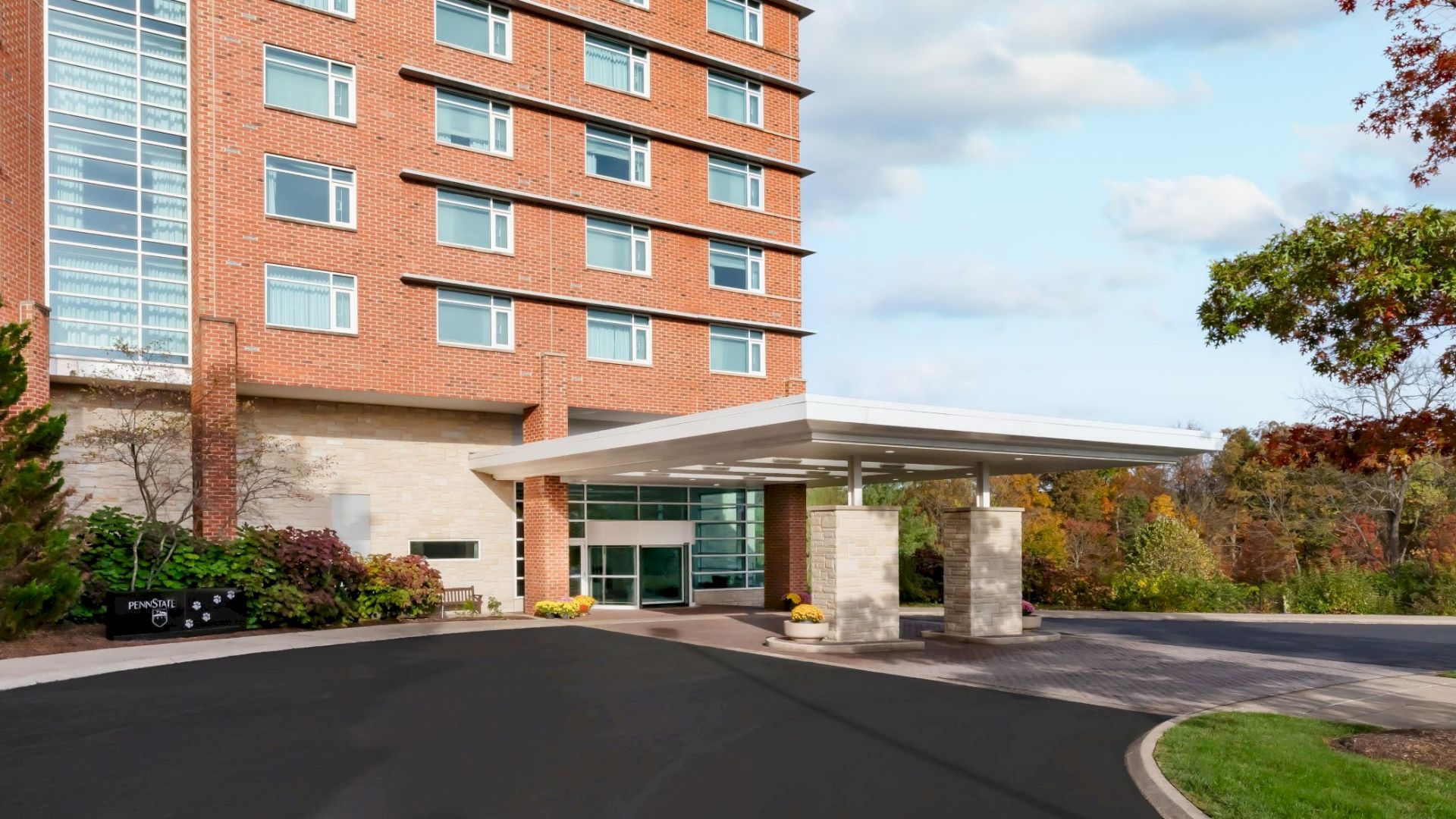 A brick hotel or apartment building with a glass atrium, driveway, and small trees; blue sky with scattered clouds, autumn foliage in the background.