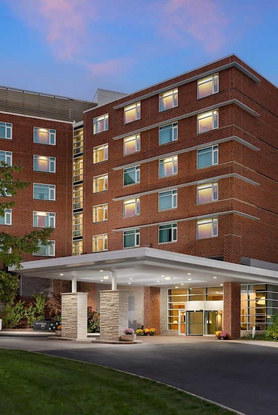 A brick hotel or dorm building with a covered entrance, multiple windows, surrounding trees, and a pink-blue sky at dusk.