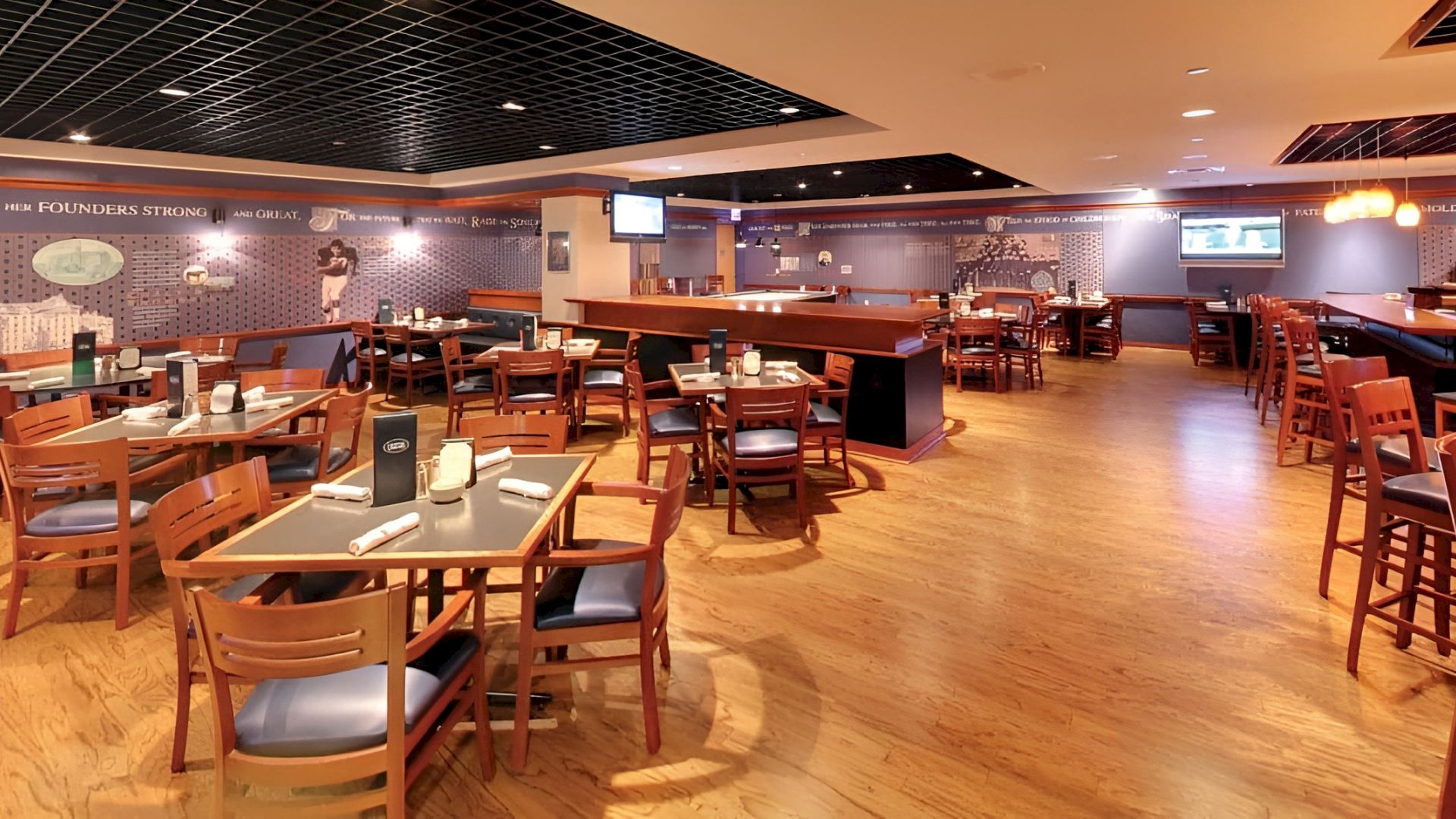 A spacious restaurant and bar interior with wooden tables, stools, warm pendant lights, and a long counter along the wall.